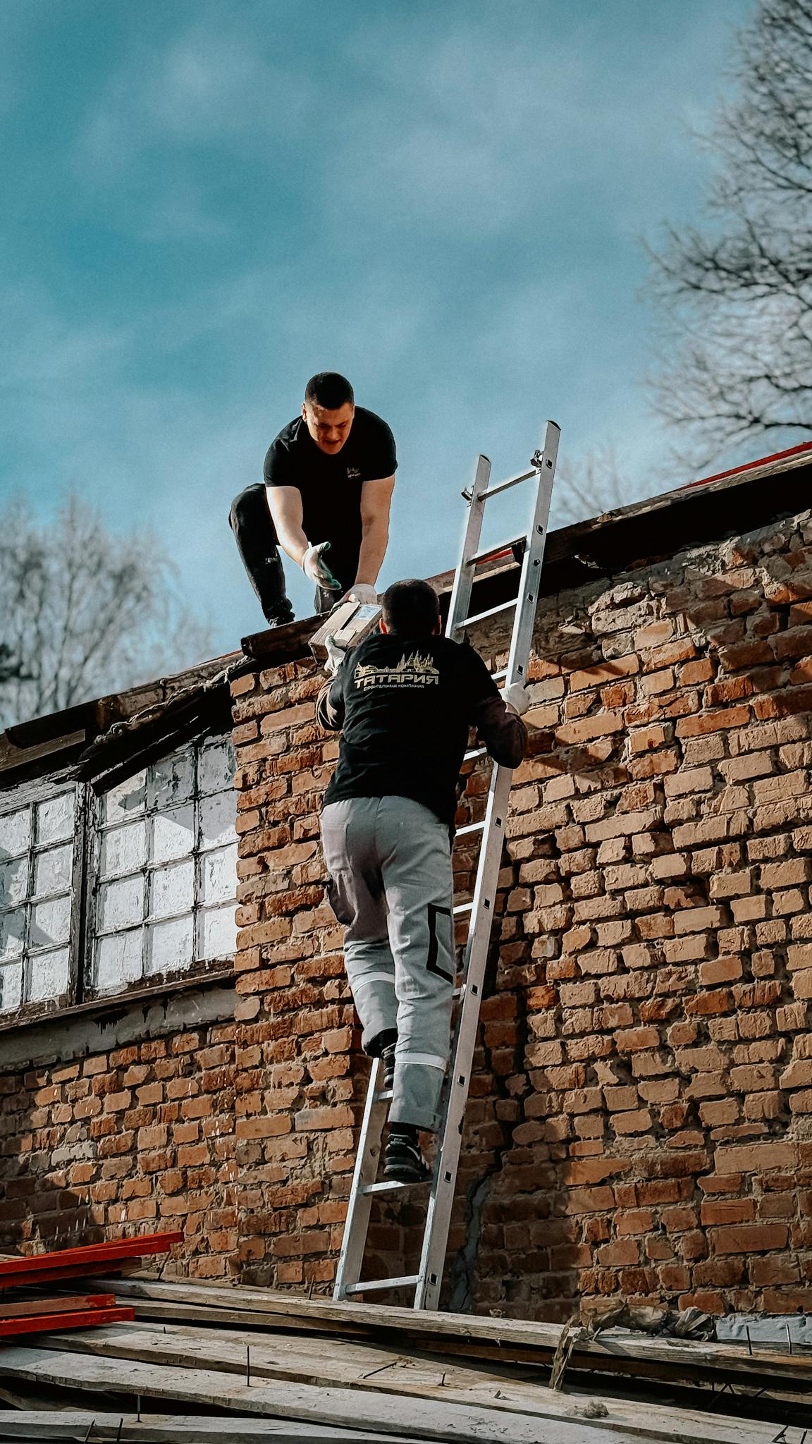 Two construction workers using a ladder to reach a brick building roof under a blue sky.