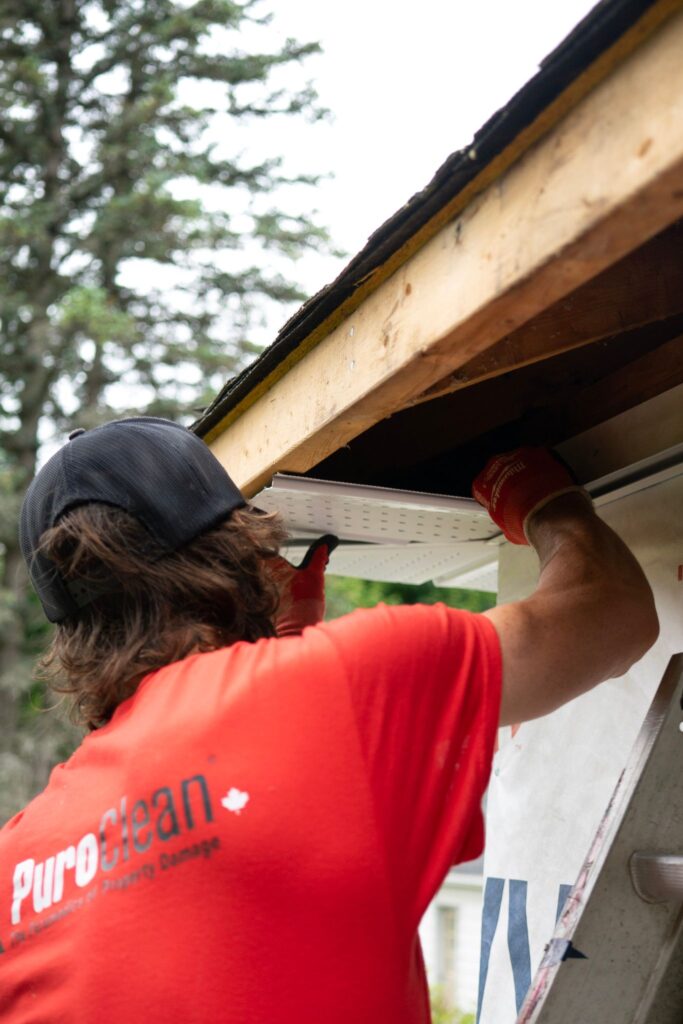 A construction worker in a red shirt installs a gutter under a wooden roof, focusing on precision.